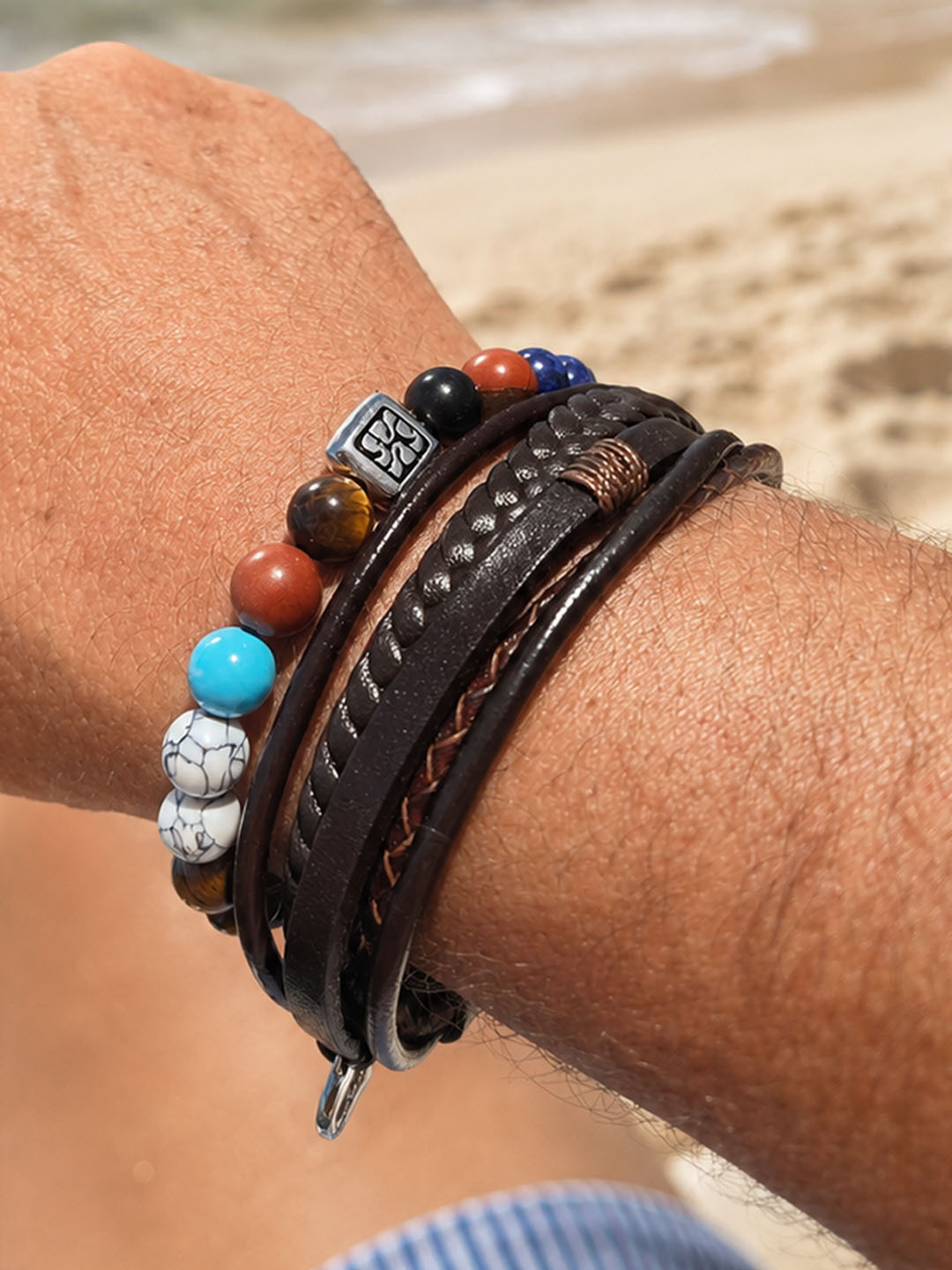 Combinatie van turquoise natuursteen kralenarmband en bruine leren herenarmband om een mannenpols op het strand.