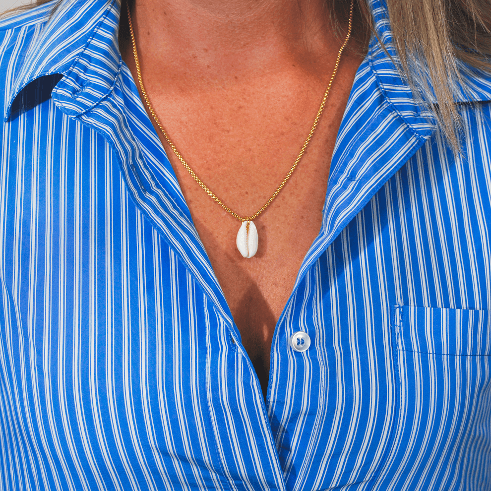 Model draagt de goudvergulde damesketting Coastline van SummerGear met witte schelphanger bij een blauw-wit gestreepte blouse.
